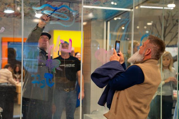 Man Taking Photograph of Friend Painting Window