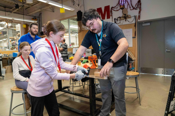 Amazeum Assistant Helps Girl Hand Saw Wood Board